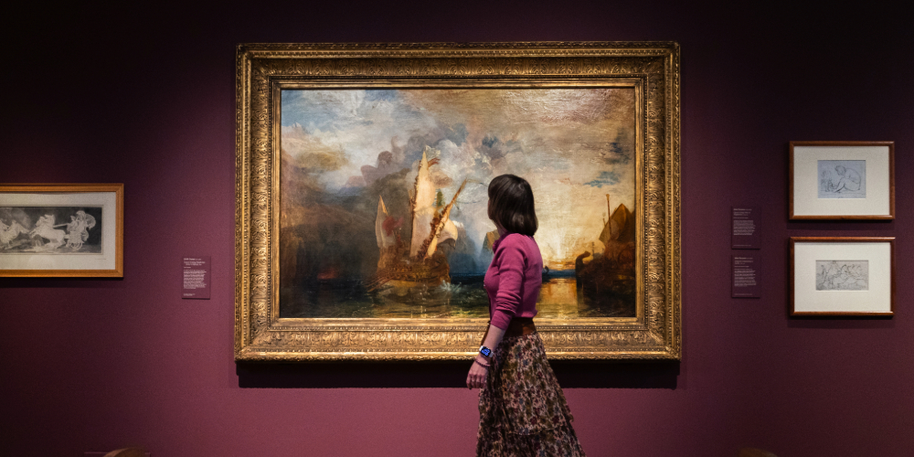 A woman walks past anoil painting of a ship at sea with cliffs and the setting sun in the background